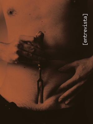 Página com uma fotografia artística em tons de sépia (marrom-avermelhado). A imagem mostra um close-up do tronco nu de uma pessoa, do peito até a cintura. As mãos seguram um garfo grande de metal com dois dentes (trinchante), posicionado verticalmente sobre o abdômen, com as pontas pressionando a região do umbigo. No lado direito, há um texto na vertical entre colchetes que diz: '[entrevista]'.